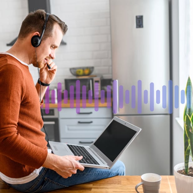 Photo of a casually dressed employee sitting on his desk working on his computer and talking into a headset.